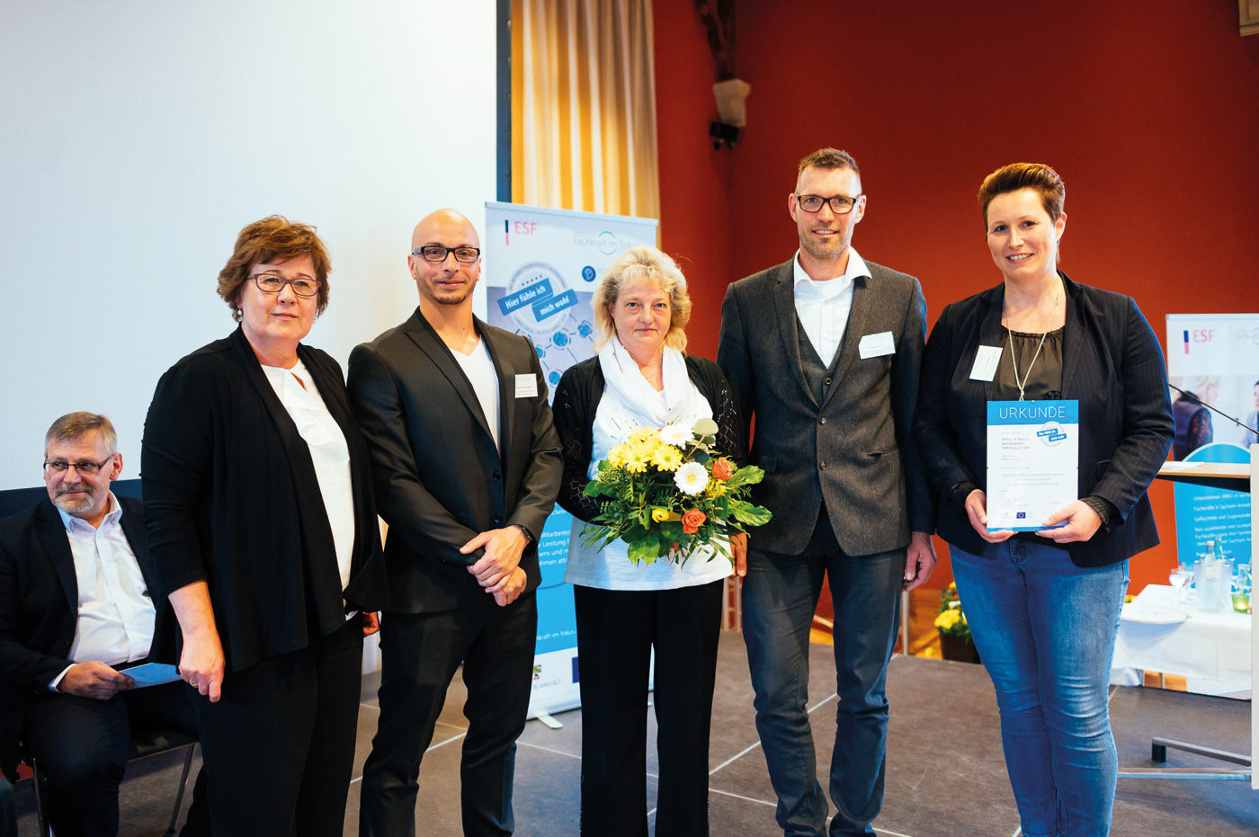 Links steht die Ministerin für Arbeit, Soziales und Integration Sachsen-Anhalt. Rechts neben ihr stehen 4 Mitarbeiter und Mitarbeiterinnen von RM Balance. Sie freuen sich und haben Blumen und eine Urkunde in der Hand. Alle schauen in die Kamera.