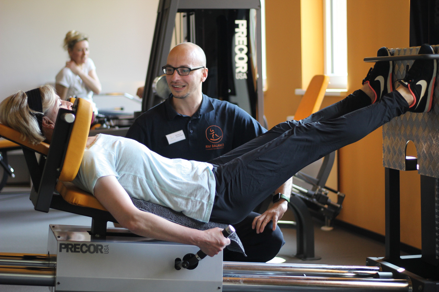 Eine Frau liegt auf einem Fitnessgerät. Im Hintergrund ist ein Mann, der sie anschaut und ihr die Fitnessübung erklärt.