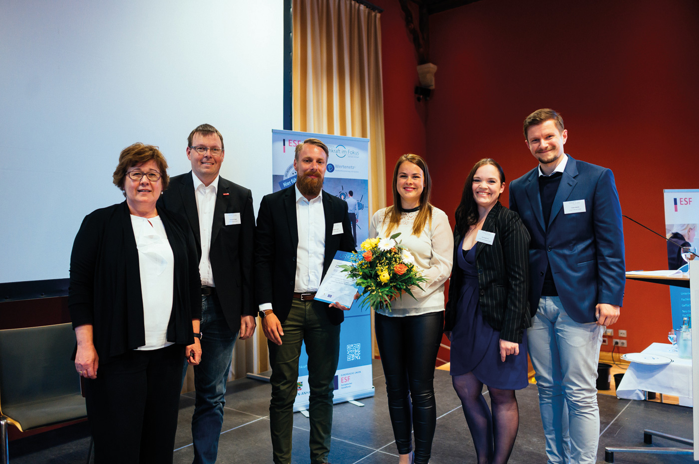 Die Minsiterin für Arbeit, Soziales und Integration steht links. Rechts neben ihr stehen 5 Mitarbeiter und Mitarbeiterinnen von Umland mit Blumen und einer Urkunde. Alle freuen sich und schauen in die Kamera.