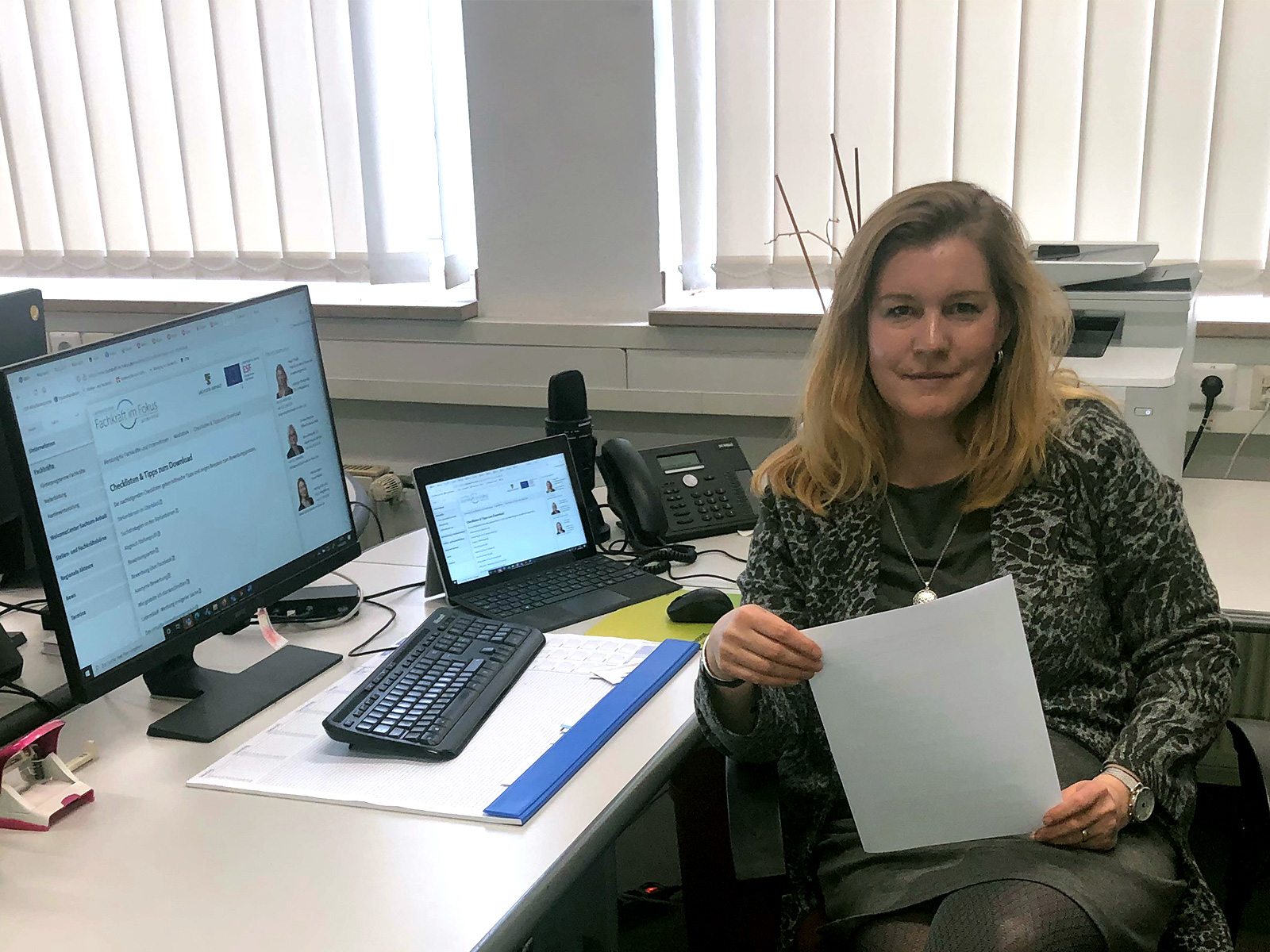 Eine Frau sitzt am Schreibtisch und hält ein Blatt Papier in der Hand
