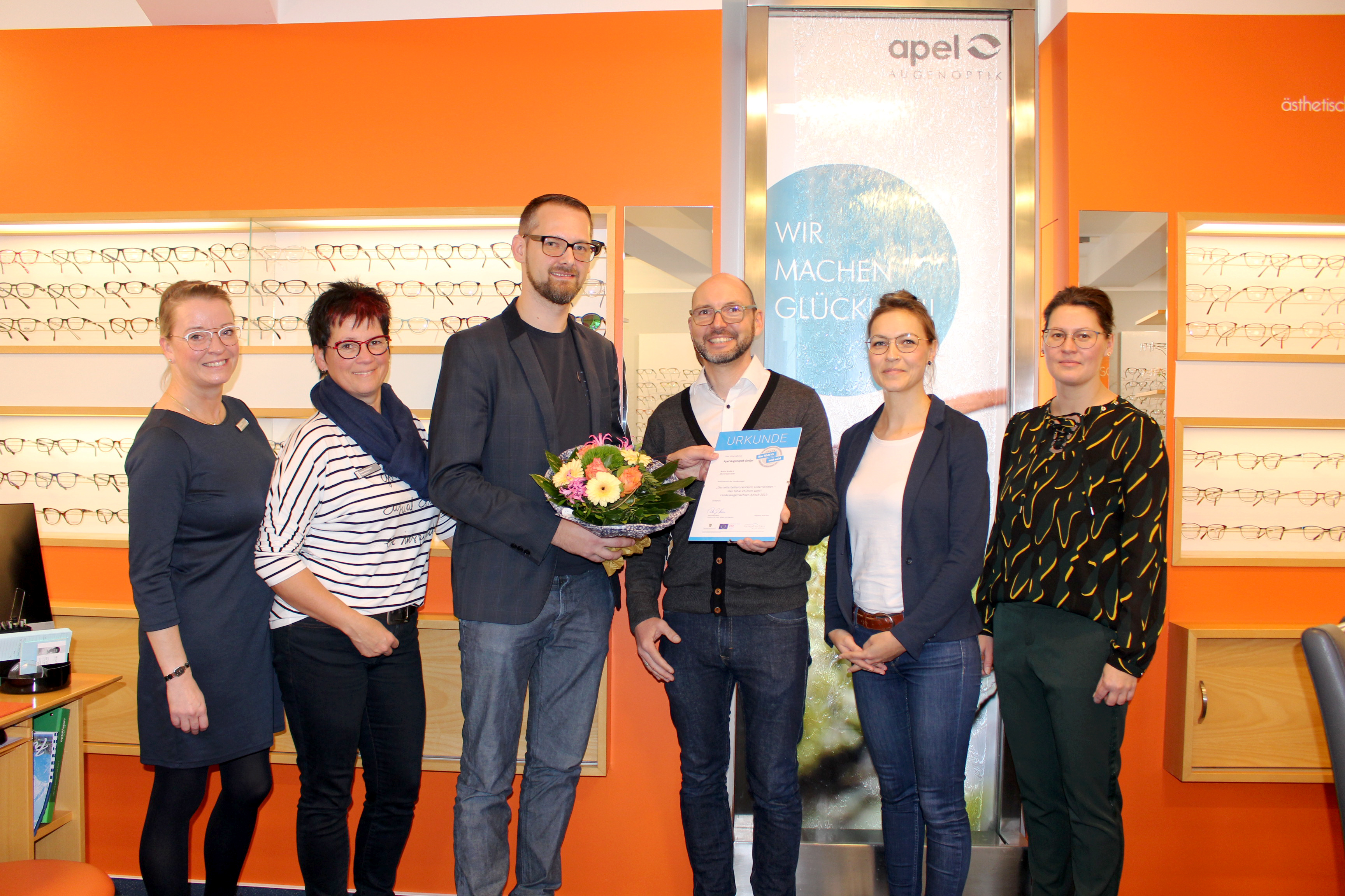 Das Team von Apel Augenoptik, 4 Frauen und ein Mann stehen vor einem Regal mit Brillen. Der Mann in der Mitte bekommt die Urkunde und einen Blumenstrauß durch Heiko Wisny der Landesinitiative überreicht. 