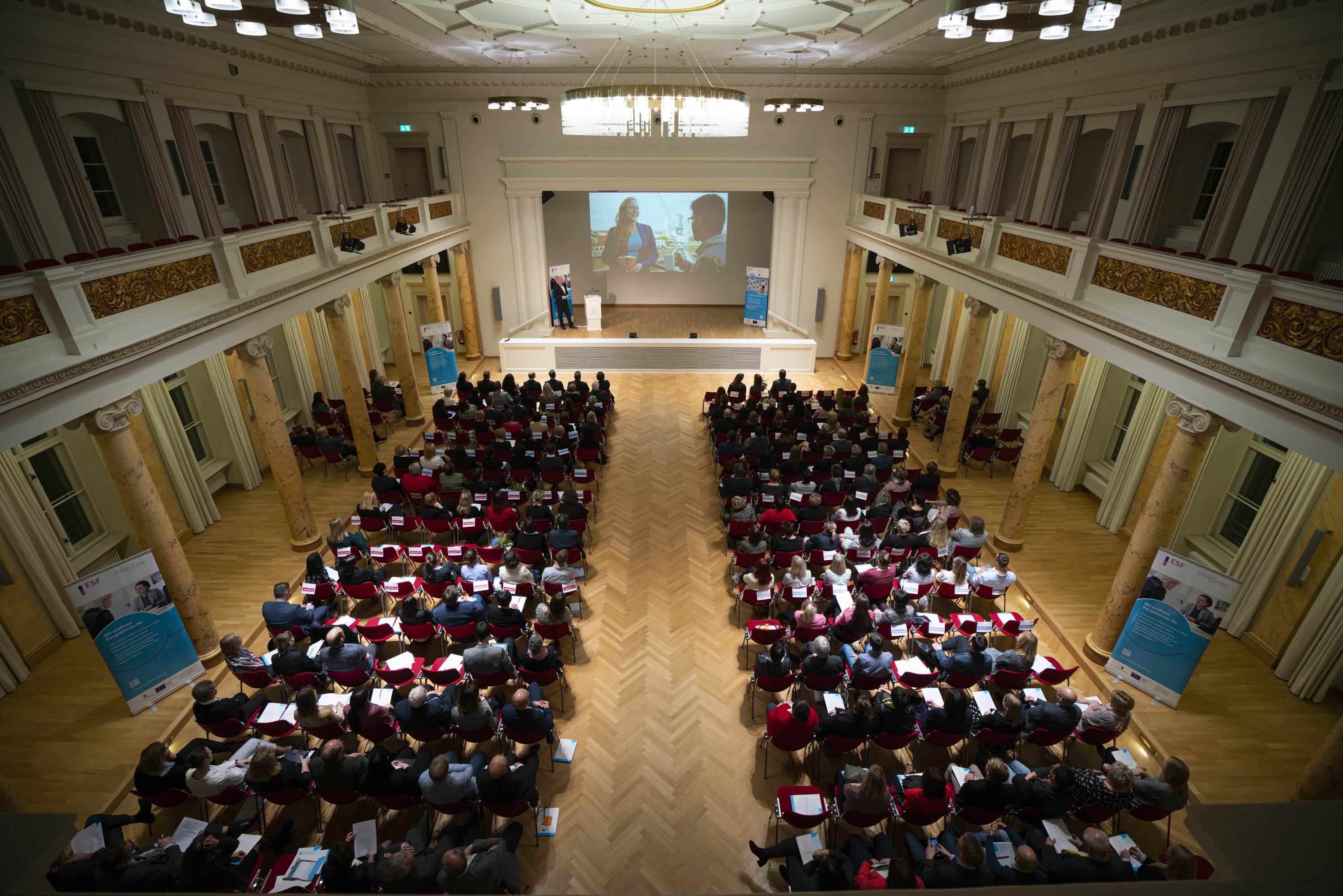 Ein Saal, vorne eine Bühne, davor viele Stuhlreihen mit Menschen 