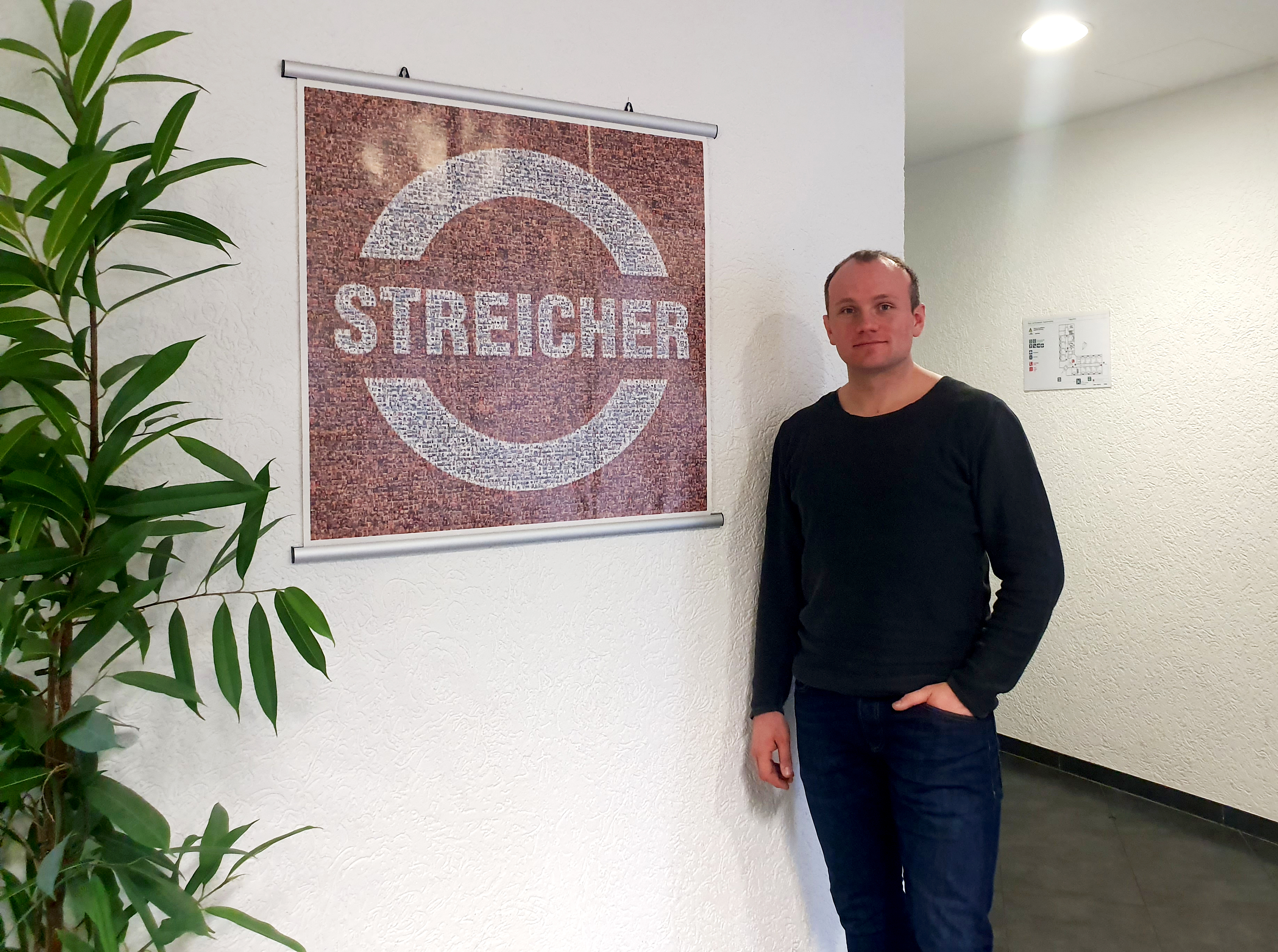 Ein Mann steht an eine weiße Wand gelehnt, links von ihm ist ein Bild auf dem steht "Streicher". Der Mann schaut in die Kamera.