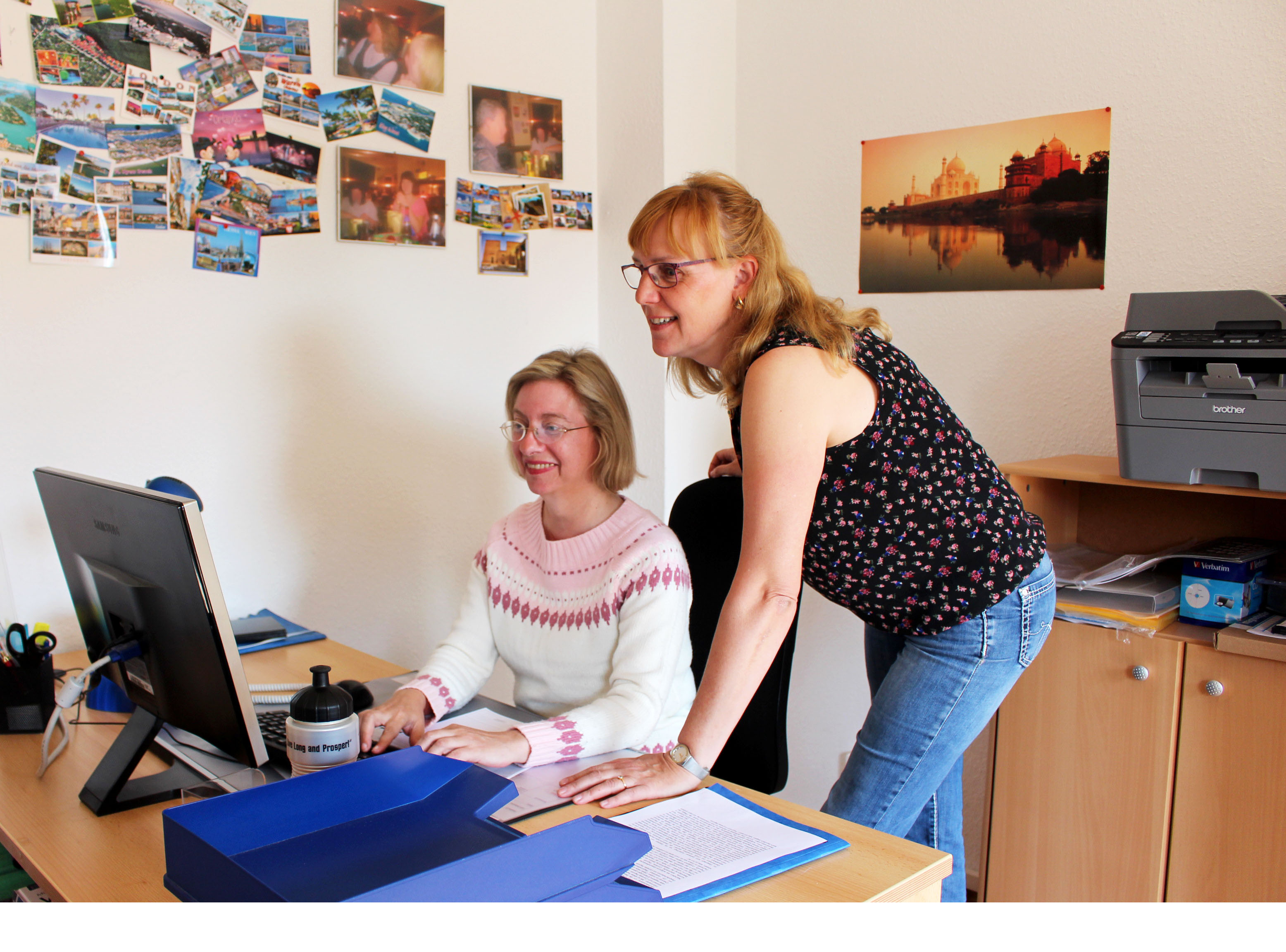 Eine Frau sitzt am Schreibtisch und arbeitet am PC. Eine zweite Frau steht neben ihr.