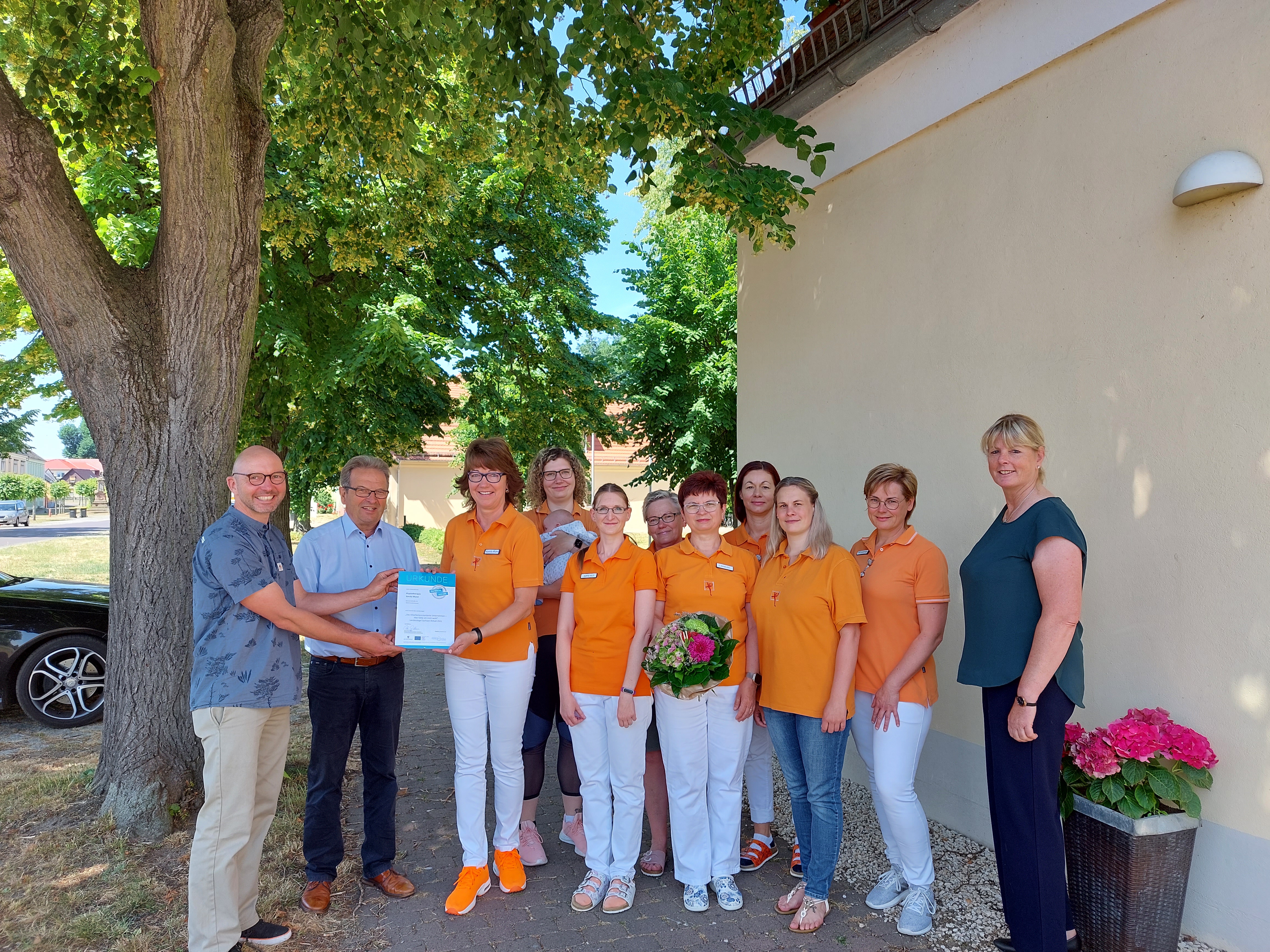 Menschengruppe steht draußen vor Gebäude, Sonne scheint, ein FRau nimmt eine Urkunde entgegen eine andere Frau hält Blumenstrauß