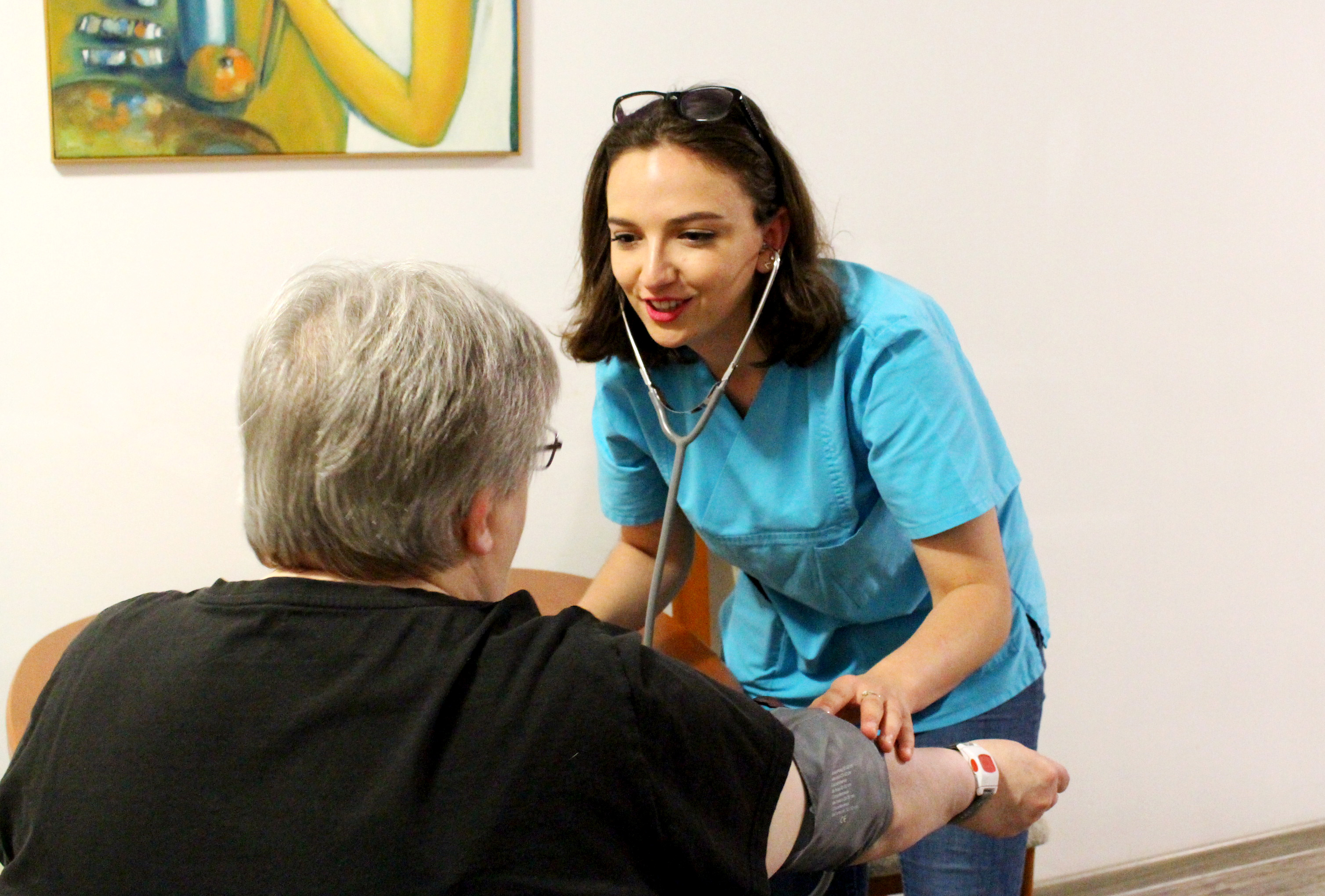 Eine Frau steht und misst einer älteren sitzenden Frau den Blutdruck.