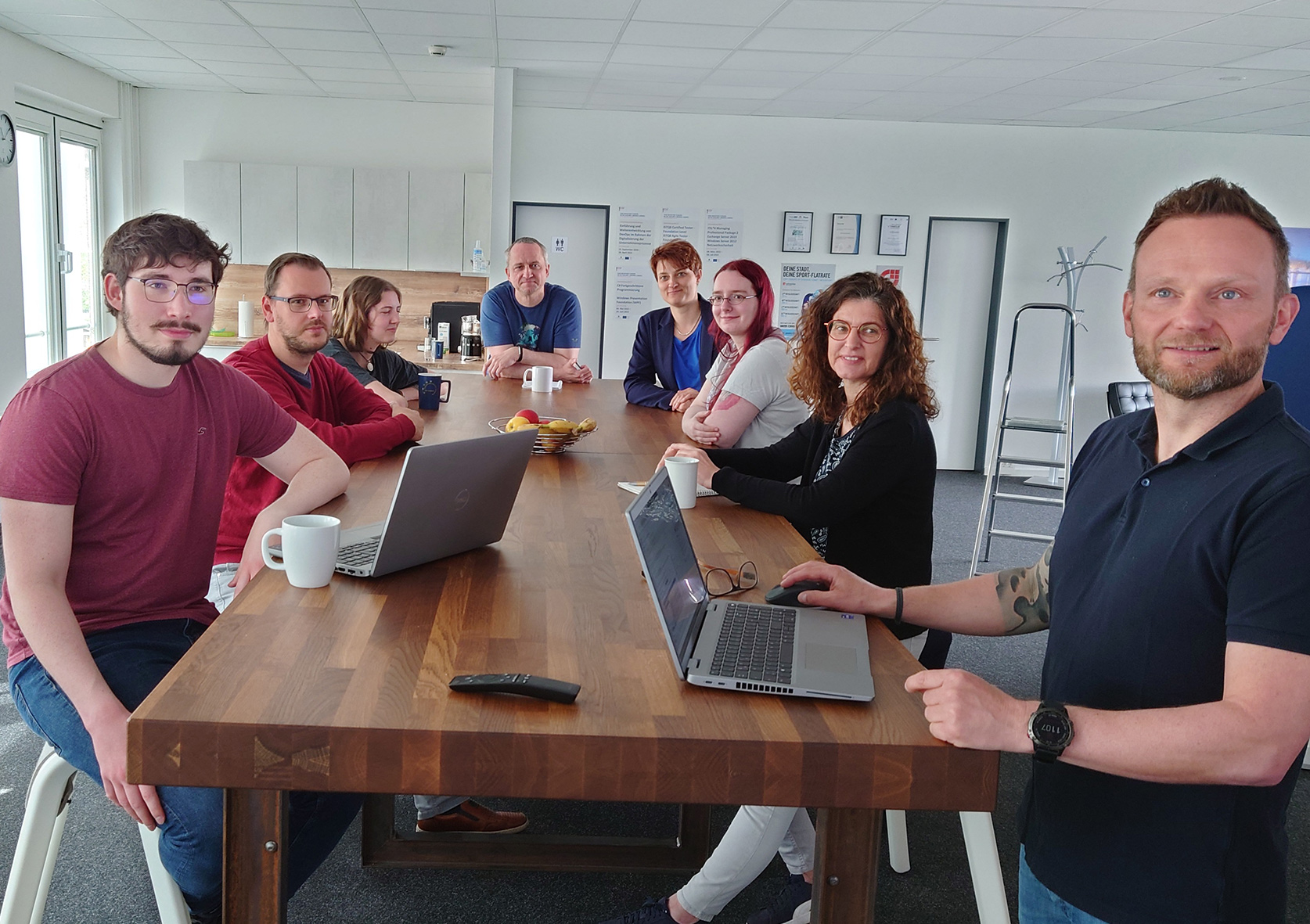 Mehrere Personen stehen an einem großen Tisch. Auf dem Tisch sind zwei Laptops und einige Tassen